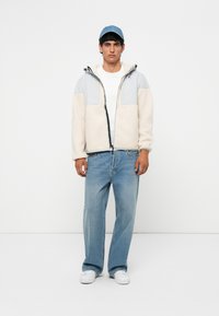 Fleece jacket in cream with gray panels, zip closure, and hood. Light blue denim cap. Light blue jeans and white sneakers complete the outfit.