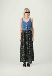 Frau trägt ein ärmelloses, blaues Denim-Hemd mit Knöpfen, einen langen schwarzen Rock mit weißen abstrakten Gesichts-Linienmustern, schwarze Sandalen mit offenen Zehen und Sonnenbrille.