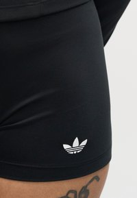 Short de sport noir avec un petit logo blanc Adidas trefoil près de l'ourlet au-dessus d'une cuisse tatouée.