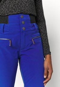 Niebieskie spodnie z wysokim stanem, zapinane na trzy czarne guziki w pasie, posiadające dwie kieszenie na zamek z złotymi akcentami oraz gładką fakturę.