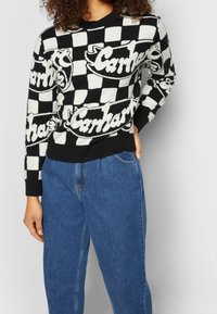 Svetr s černobílým kostkovaným vzorem obsahující velké logo "Carmar". Vyrobeno z pleteného materiálu, s kulatým výstřihem a žebrovanými manžetami.