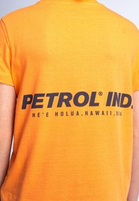 Camiseta de algodón naranja con un atrevido estampado de texto negro en la parte posterior, que dice "PETROL IND. HE'E HOLUA, HAWAI, EE. UU." Manga corta.