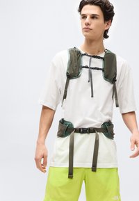 Νεαρός άνδρας που φοράει λευκό μπλουζάκι και κοντό παντελόνι νέον κίτρινου χρώματος με εξάρτηση τύπου ιμάντα πράσινου χρώματος, που έχει αγκράφες και ιμάντες στους ώμους και τη μέση.