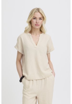 Femme blonde portant un haut crème à manches courtes et col en V, ainsi qu'un pantalon assorti, debout les mains dans les poches devant un fond uni.