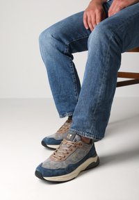 Le sneakers presentano un mix di materiali in suede grigi e blu, con una superficie testurizzata, suola beige e lacci marroni. Dettaglio del logo dorato sul lato.