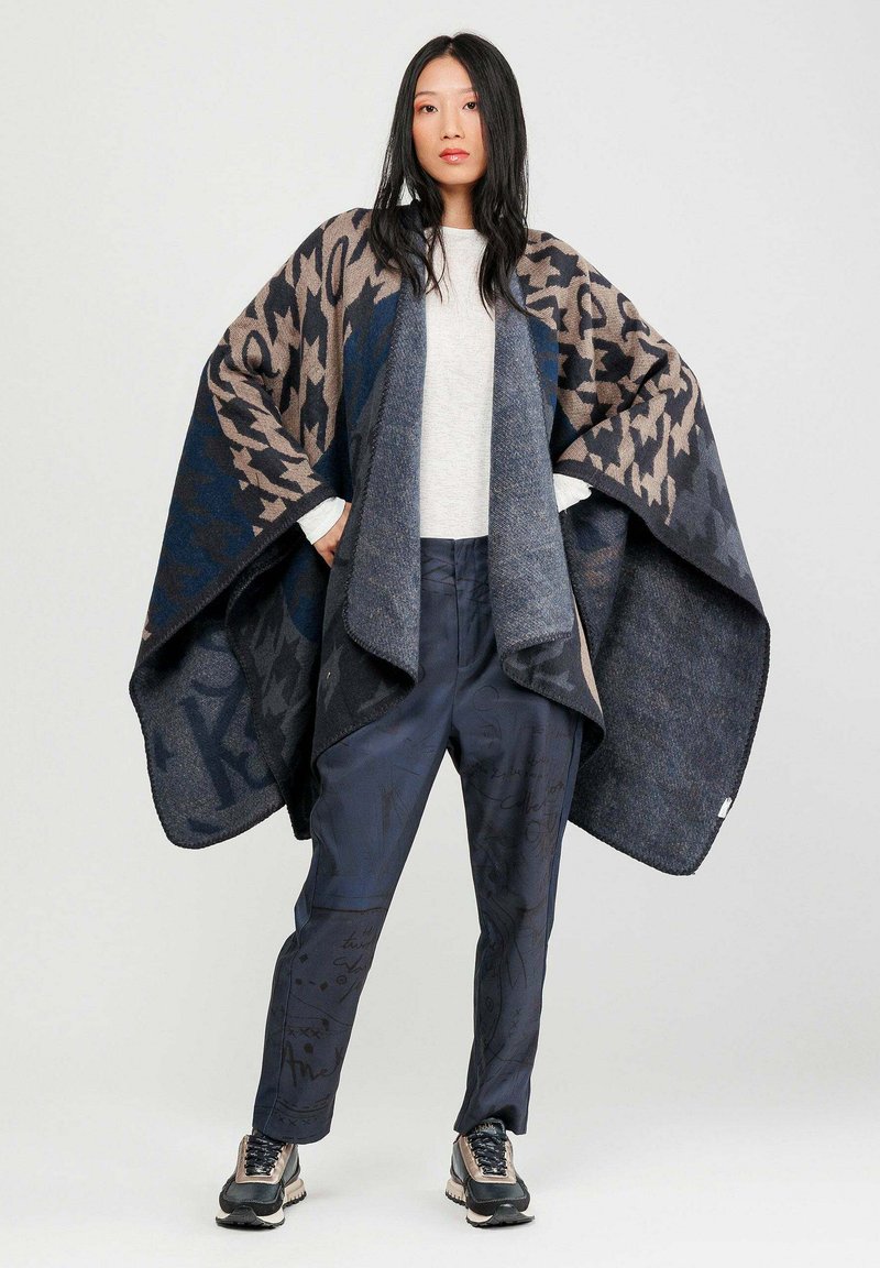 Poncho de lana mezcla de gran tamaño en patrones geométricos azul marino y marrón, usado sobre una blusa de manga larga clara y pantalones estampados en azul marino.