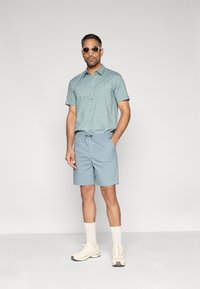 Ανοιχτό μπλε πουκάμισο με κοντά μανίκια και μικρές λευκές βούλες, συνδυασμένο με ταίριαστο ανοιχτό μπλε σορτς. Λευκά αθλητικά παπούτσια και γυαλιά ηλίου ολοκληρώνουν την εμφάνιση.