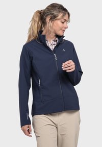 Marinefarbener Softshell-Jacke mit hohem Kragen, Reißverschluss-Taschen auf der Vorderseite und Logo auf der Brust. Glatte Textur, schlanke Passform, getragen über einem karrierten Hemd.