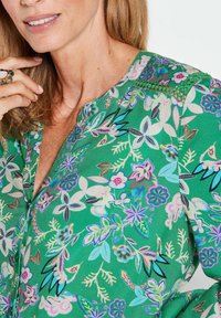 Groene bloemenblouse met een V-hals en korte mouwen, versierd met verschillende pastelgekleurde bloemen en bladeren in een herhalend patroon.