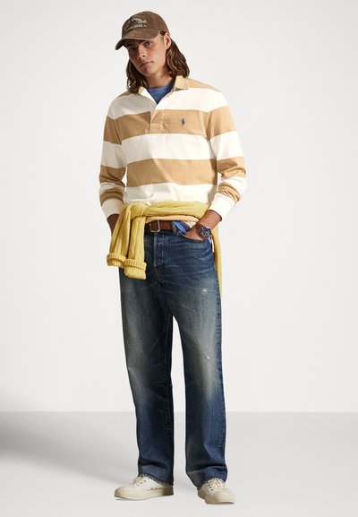 Jauns vīrietis valkā bēša un baltas svītrains regbija kreklu, zilas džinsas, bēša cepuri, baltus sporta apavu un dzeltenu džemperi, kas sasiets ap vidukli, stāv ar rokām kabatās.