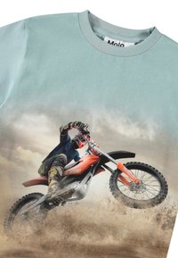 Camiseta de algodón azul claro con un gráfico de un piloto de motocross en acción, con salpicaduras de tierra y una posición dinámica.