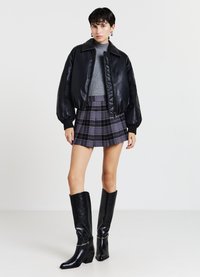 Giubbotto bomber in pelle nera con colletto alto, maglione aderente grigio, gonnellina a quadri in tonalità di viola e stivali neri fino al ginocchio.