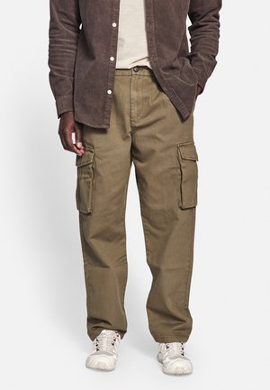 Mann trägt braune Cargohosen, weiße Sneakers, cremefarbenes Hemd und ein braunes Hemd mit Knöpfen, Hände in den Taschen.