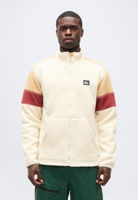 Άνδρας με κρεμ φλις μπουφάν με μπεζ και κόκκινες ρίγες στα μανίκια και πράσινο παντελόνι, στέκεται μπροστά σε ένα απλό φωτεινό φόντο.