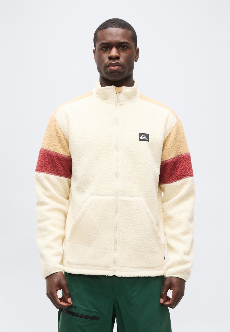 Άνδρας με κρεμ φλις μπουφάν με μπεζ και κόκκινες ρίγες στα μανίκια και πράσινο παντελόνι, στέκεται μπροστά σε ένα απλό φωτεινό φόντο.