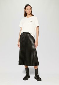 T-shirt blanc à manches courtes avec "bead" brodé en marron, associé à une jupe midi plissée noire et des bottes de cheville noires chunky.