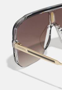 Γυαλιά ηλίου Carrera με μαύρο και διαφανές πλαίσιο από πλαστικό. Διαθέτουν καφέ φακούς με κλίση και χρυσές λεπτομέρειες στα βραχίονες με μαύρες ρίγες.