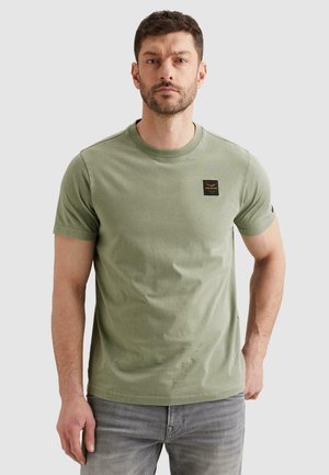 Mann trägt ein olivgrünes kurzärmeliges T-Shirt mit einem kleinen schwarzen Fleck auf der Brust, kombiniert mit hellgrauen Jeans, steht neutral.