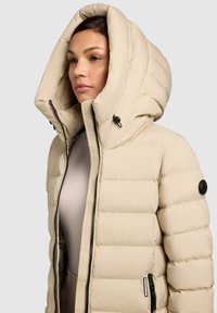 Valge-beež värvi puffer-jope suure kapuutsi, tikitud mustriga, kahe tõmblukuga tasku ja varrukal detailina logonupuga.