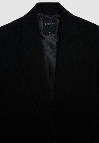 Blazer nero con finitura liscia, caratterizzato da sottili perforazioni e interno in raso. Etichetta con la scritta "CALLIOPE," taglia S. Design classico e sartoriale.