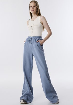 Pantalones de chándal de pierna ancha en azul claro, con bolsillos laterales, cinturilla elástica y tachuelas decorativas a lo largo del costado. Combinados con una camiseta de tirantes crema.