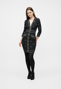 Γυναίκα που φοράει εφαρμοστό μαύρο φόρεμα με γεωμετρικό σχέδιο, χρυσά κουμπιά, μανίκια τριών τετάρτων, μαύρο καλσόν και μαύρες ψηλοτάκουνες, όρθια.