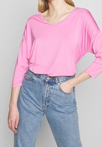 Maglietta rosa a maniche lunghe con scollo a V abbinata a jeans di denim azzurro chiaro a vita alta. La maglietta ha una vestibilità comoda con maniche drappeggiate.