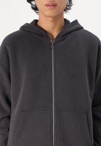 Donkergrijze zip-up hoodie met een zachte textuur, kangaroo zakken en een eenvoudig ontwerp. Beschikt over een grote capuchon en een zilveren rits.
