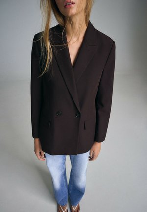 Femme portant un blazer noir croisé, un jean bleu clair délavé et des tongs blanches, debout devant un fond gris uni.