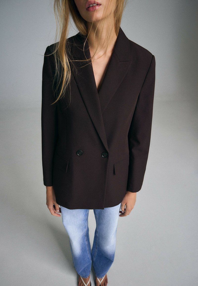 Femme portant un blazer noir croisé, un jean bleu clair délavé et des tongs blanches, debout devant un fond gris uni.