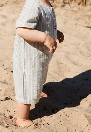 Combinaison légère rayée en bleu pâle et blanc, manches courtes, fermeture à boutons, portée par un enfant debout pieds nus sur un sol sablonneux.