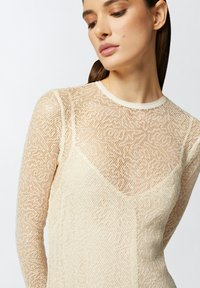 Pleteni dugi rukav u krem boji, s mrežastim uzorkom, okruglim izrezom i uskim siluetom. Tekstura izgleda lagano i prozirno.