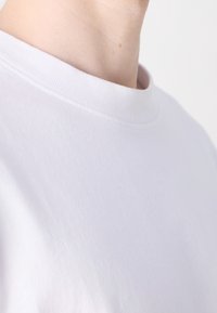 Gros plan sur une personne portant un t-shirt blanc uni à col rond, montrant la région du cou et des épaules sur un fond blanc.