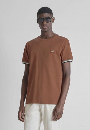 Jonge man met grote zwarte zonnebril, bruin kortärmelig shirt met klein logo en witte broek, staand tegen een lichte effen achtergrond.