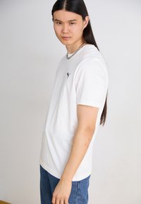 Hvit kortermet t-skjorte med svart logo på venstre bryst, kombinert med blå denimjeans. Stoffet virker glatt med en avslappet passform.