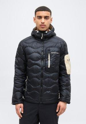 Jeune homme portant une veste matelassée noire avec une fermeture éclair frontale, une capuche, une poche poitrine et un patch beige sur la manche gauche, debout devant un arrière-plan uni.