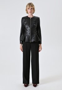Giacca peplum nera in pelle con collo alto, corpo aderente e chiusura con zip dorata, abbinata a pantaloni neri a gamba larga.