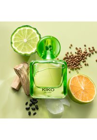 Grønn glassparfymeflaske med rund kork, omgitt av skivet lime, appelsin, korianderfrø, tre og klart krystall på en lys grønn bakgrunn.