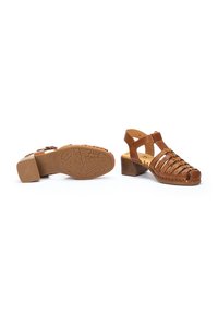 Des sandales à talons en cuir brun présentant des brides avec des découpes, un bout ouvert et une semelle en caoutchouc texturé avec un motif géométrique.