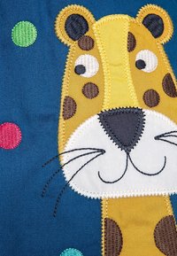 Żółto-brązowy haftowany wizerunek lwa na niebieskim tle, z zielonymi i czerwonymi kropkami. Miękka tekstura, wyraźne cechy.