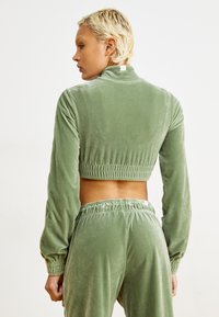 Chándal de terciopelo verde que incluye una chaqueta corta con cierre de cremallera, de mangas largas y cintura elástica, acompañado de pantalones a juego. Textura suave.