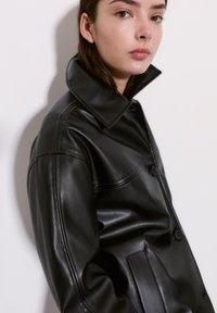 Czarna skórzana kurtka z wysokim kołnierzem, zapinana na guziki, z bocznymi kieszeniami. Gładka faktura z lekkim połyskiem i minimalnymi przeszyciami.