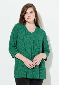 Zielona bluzka z dekoltem w serek, z teksturowym wzorem małych, ciemnozielonych kształtów, o rękawach długości 3/4 i luźnym kroju, zestawiona z czarnymi spodniami.