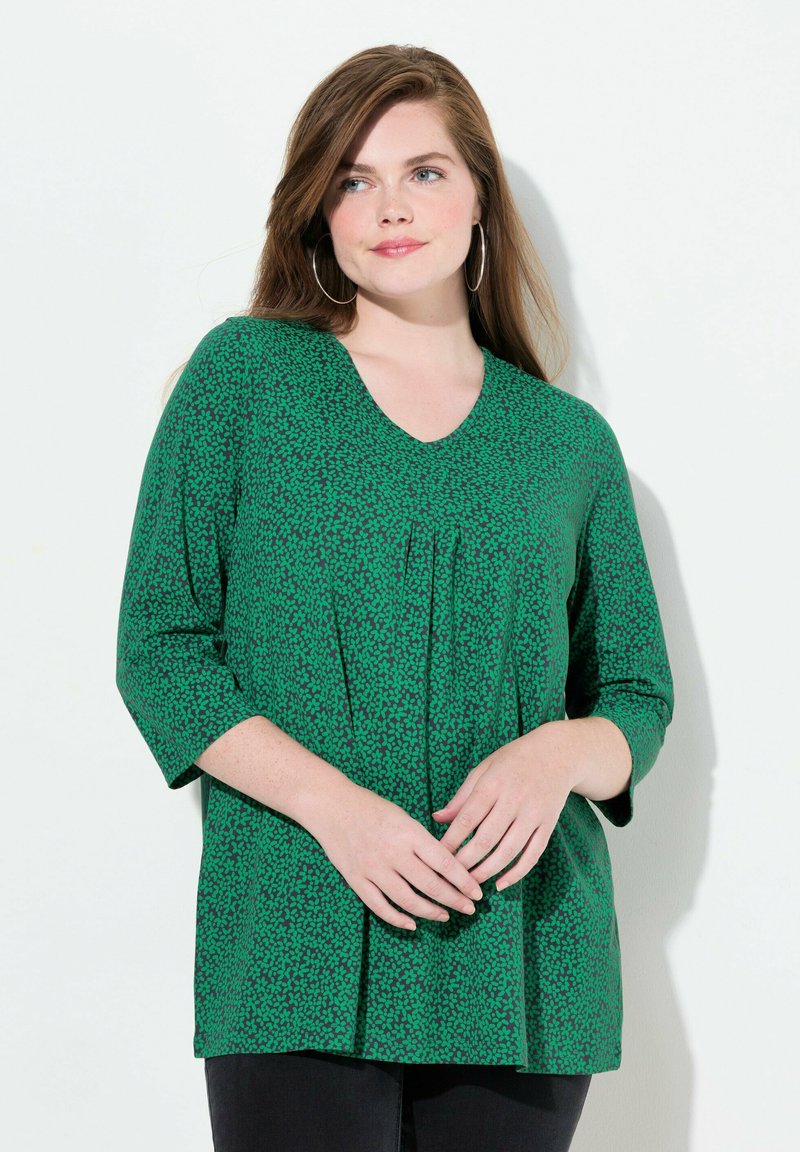 Zielona bluzka z dekoltem w serek, z teksturowym wzorem małych, ciemnozielonych kształtów, o rękawach długości 3/4 i luźnym kroju, zestawiona z czarnymi spodniami.