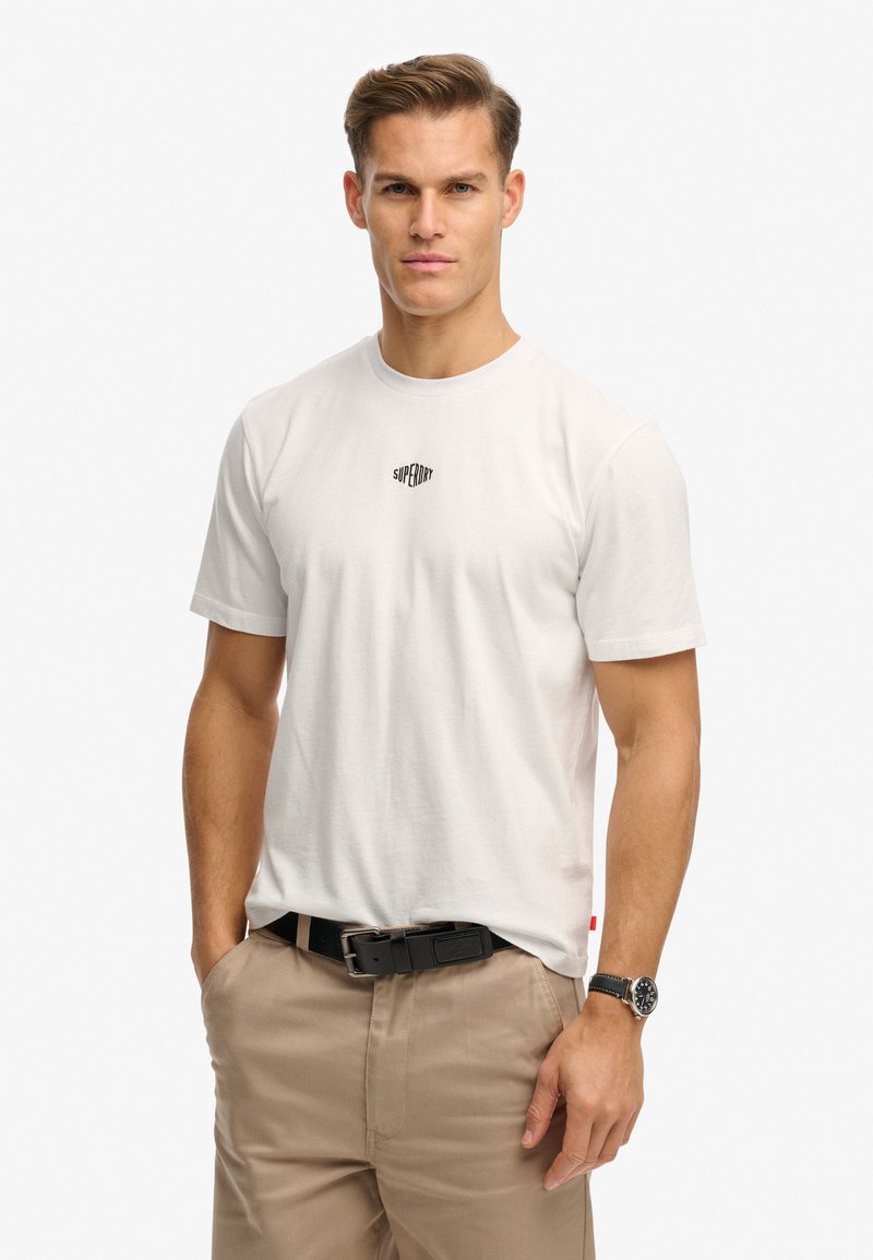 Homme portant un t-shirt blanc Superdry, un pantalon beige avec une ceinture noire et une montre-bracelet noire, debout avec une main dans la poche.