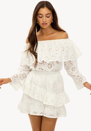 Mujer con vestido blanco de ojales sin hombros con cortes florales, capas con volantes, mangas largas y pendientes de aro dorados, mirando hacia abajo con los brazos ligeramente extendidos.