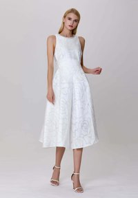 Femme en robe midi sans manches blanche à motif floral et sandales à talons blancs, se tenant devant un fond gris uni.