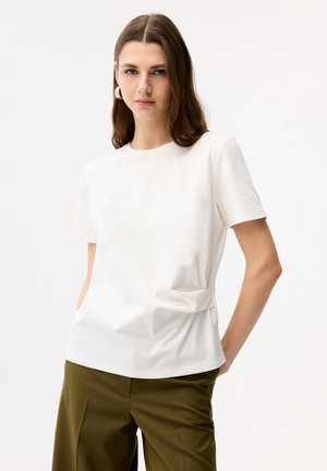 Junge Frau mit langen braunen Haaren, die ein weißes kurzärmliges Oberteil und olivgrüne Hose trägt, mit den Händen in den Taschen stehend.