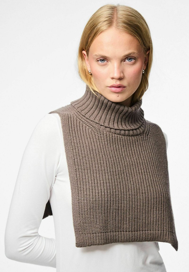 Ruskea neulottu kauluspaidalla, jossa on teksturoitu, ribattu kuvio. Hihaton malli kerrostettuna valkoisen pitkähihaisen paidan päälle.