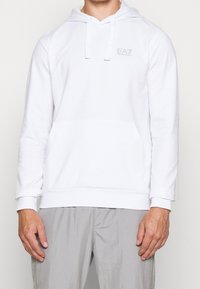 Bílá mikina s kapucí z měkké látky, vybavená přední kapsou ve tvaru klokánky a jemným logem EA7 na hrudi. Dlouhé rukávy s žebrovanými manžetami.